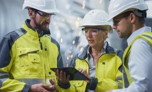 A woman holding a tablet speaks to two men. They are wearing high visibility jackets and hard hats. Sparks can be seen in the background coming from a piece of machinery.