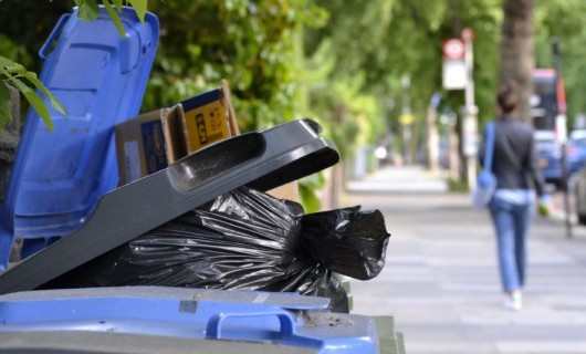 A close up of several wheelie bins over flowing with the amount of black bin bags inside them. A woman walks down the street in the distance beside a line of parked cars.