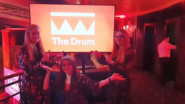 Savannah, Daisy and Jo posing and pointing to a screen displaying The Drum logo.