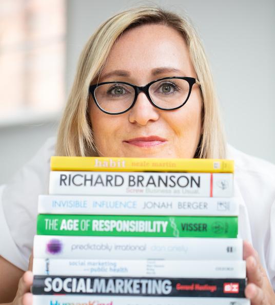 Chief Executive, Kelly, rests her chin on a pile of books in front of her, smiling.