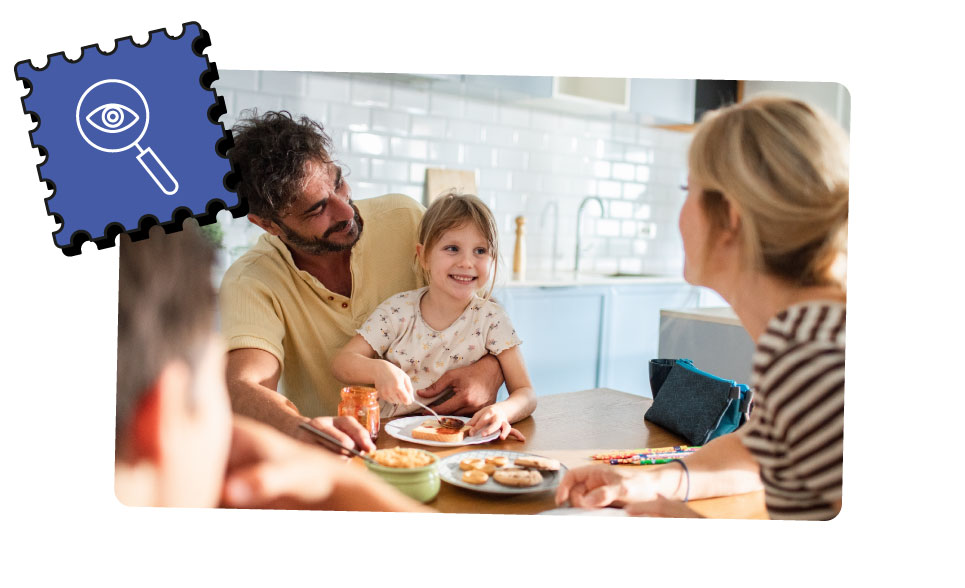 A family of 4 eat breakfast together around the table. A young girl sitting on her fathers lap also spreads jam on her toast.